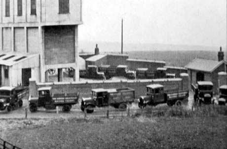 Photograph showing eight motor lorries driving one behind the other round a large concrete building, through a gap in the wall and along a road outside the wall; the lorries have the words The Horden Collieries Ltd. Blackhall Colliery on them; they have been identified as First Coal Lorries, Blackhall