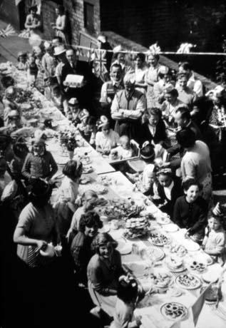 Photograph showing a long table, running the length of the photograph from the bottom right-hand corner to the top left-hand corner, with plates of food on it; children are sitting either side of it and adults are standing behind them watching the proceedings; Union Jacks are strung over the table; the photograph has been described as Meadow Avenue Street Party, Blackhall; the occasion for the street party is not clear and it may be a party to celebrate the end of World War Two or the Coronation of Queen Elizabeth II