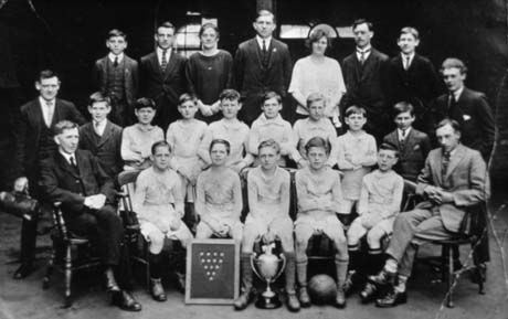 Photograph of eleven boys, aged approximately eleven years, in football strip, posed with four other boys, eight men and two women; in front of the group is a trophy cup and a board on which there are fourteen small shields; the group has been described as Blackhall Granary Football Team