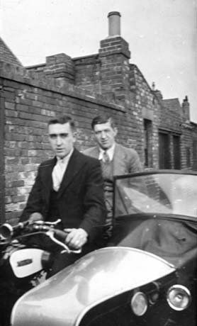 Photograph showing two young men, one of whom has been identified as J. Barclay, sitting on a motor bicycle; the handle bars of the bicycle and the bonnet of the side car can be seen; both men are dressed in suit and tie; behind them are the back walls of the yards of terraced houses identified as being in Blackhall