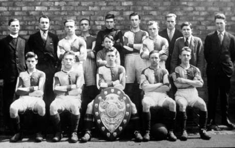 Photograph of ten young men and one boy, aged approximately thirteen years, posed against a brick wall, accompanied by four other men and a clergyman; a large shield is on the floor in front of the front row; they have been identified as Blackhall, St. Andrew's, Football Team