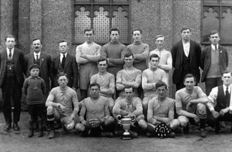 Photograph of twelve men in football strip, posed in front of a building with painted glass windows, accompanied by six other men and a boy, aged approximately ten years; in front of the group, is a trophy cup, a board with medals on it, and a football, possibly reading: Blackhall Wesleyan Athletic Football Club; the photograph has been identified as Wesleyan Cup, Blackhall
