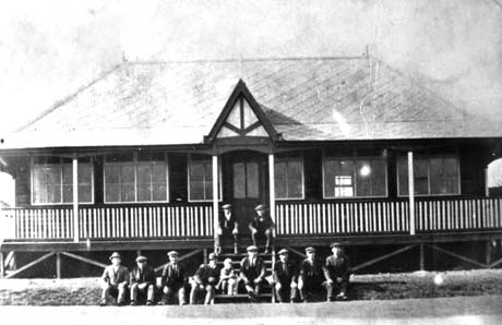 Photograph of ten men wearing suits and caps sitting on the steps leading up to a pavilion, a single-storey building with a veranda; a small girl and a dog is also sitting on the steps; the whole of the front of the pavilion can be seen; the photograph has been identified as Blackhall Bowls