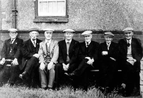 Photograph of seven elderly men wearing suits and caps sitting in a row against the wall of a house; they have been identified as Mr. Jack Parker and Friends