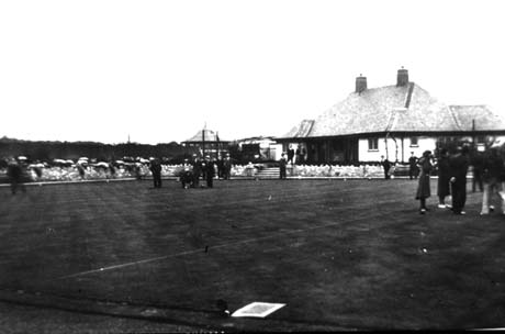 Photograph showing the surface of a grassed open space surrounded with flower beds and with a single-storeyed building on the right; indistinct figures can be seen standing on the grass; the photograph has been identified as Blackhall Bowls