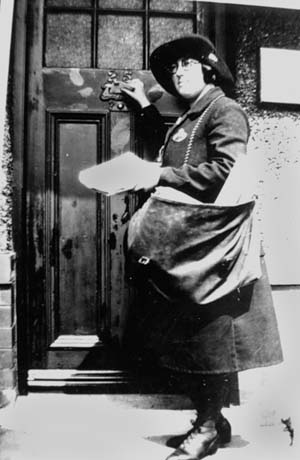 Photograph showing a woman wearing a coat, an ankle-length skirt, boots and a hat, standing in front of a door with her hand on its knocker; she is also carrying a large bag over her left shoulder; she has been identified as Connie Naylor, Post Girl of Blackhall