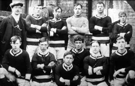 Photograph of eleven young men in football strip posed in the backyard of a house, showing a window with a lace curtains, a tin bath on the wall and a dividing wall between two yards; the young men are accompanied by a man in a suit and cap; they have been identified as Blackhall United First Football Team
