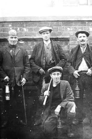 Photograph showing four men posed against a brick wall; the man on the left of the picture is wearing a suit, and shirt and a leather cap on his head and is carrying a miner's lamp and a stick; the two younger men standing by him against the wall are wearing jackets, shirts, waistcoats and caps and are carrying miner's lamps; the man kneeling in front of the others is dressed in a similar manner to the two other young men, but is carrying a pick over his right shoulder; they have been identified as Blackhall Miners