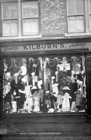 Photograph of the window of a draper's shop showing the contents of the window and a sign reading Merry Xmas at the front of the window; the sign above the shop reads: Drapers Kilburn's Milliners; three windows on the floor above have signs reading: Corsets, Blouses, and Baby Linen; the shop has been identified as being in Blackhall