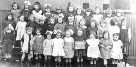 Photograph of a group of thirty three small girls, aged approximately six years, posed against the wall of a corrugated iron building; they have been identified as pupils at Blackhall Council Infants' School