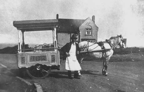 Photograph of a man wearing a jacket, tie, long apron and a hat, standing in front of the shafts of a cart in which a horse is harnessed; the cart has two wheels partly covered by a wooden structure on which the words, B. Pieroni and Son, are written; the cart is also covered by a canopy; behind the horse and cart an isolated detached house cab be seen