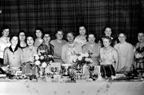 Photograph of fourteen women, dressed in formal day dresses, standing behind a table on which trophies,flowers and handbags can be seen; the photograph has been identified as Ladies Bowls Presentation, Blackhall