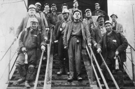 Photograph showing fourteen coal miners walking down steps towards the camera; they are wearing hard hats, carrying miner's lamps and have coal dust on their faces; they can be seen full length and close-up; they have been identified as miners at Blackhall Colliery