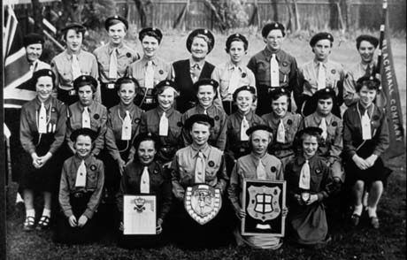 Photograph of twenty one girls, aged between eleven years and fifteen years, dressed in the uniform of the Girl Guides, posed in a garden; a woman dressed as a Guider is on the back row; on the left is a Union Jack and on the right a flag on which the words Blackhall Company can be seen; three girls on the front row are holding a certificate, a shield, and a coat of arms