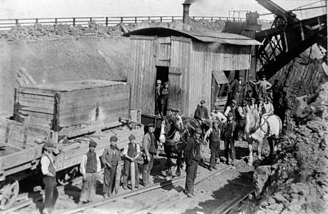 Photograph showing fifteen workmen standing along a railway line in front of a railway waggon with a wooden box on it and a railway waggon with a corrugated iron hut with a smoking chimney on it; two further men can be seen at the doorway of the hut and three men are at its side; a horse is standing near the line; the railway is in a cutting and the sides of the cutting can be seen; part of a bridge can be seen at the right of the picture; the photograph has been described as Building The Coast Railway, Blackhall