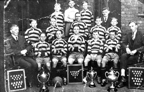 Photograph of thirteen boys, aged approximately fourteen years, posed with three men in front of a brick house; in front of the group are three trophy cups and three boards containing medals; they have been identified as Blackhall Football Team