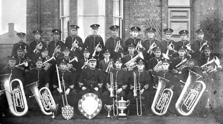 Photograph of twenty eight men in uniform, carrying musical instruments, posed in front of the bay window of a house; in front of the group are three trophy cups, a shield and a shield on which there are medals; they have been identified as members of the Blackhall Colliery Band
