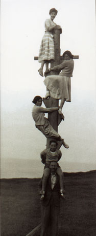 Photograph showing three young women dressed in skirts and cardigans standing on foot rests on a crucifix-shaped pole; at the bottom of the pole is a man with a child on his shoulders; an indistinct landscape can be seen behind the pole, which has been identified as being in Blackhall