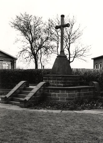 Photograph of a stone cross with a cone-shaped base on which the names of those killed in battle are inscribed; below the cross are two walls and a flight of steps to reach the base of the memorial on top of the walls; in the distance are a hedge and the windows and roofs of two low buildings; it has been identified as being in Blackhall