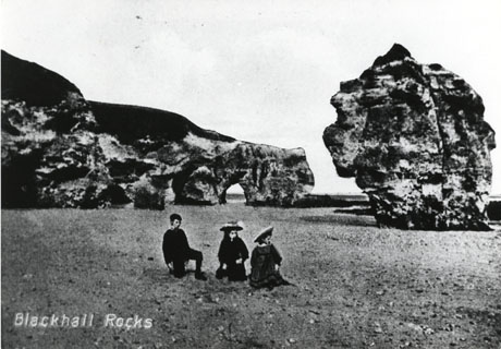 Postcard photograph entitled Blackhall Rocks, showing part of the cliff extending along the beach with a hole in it ; an individual rock can also be seen; a small boy and two small girls can be seen sitting on the beach in the forefront of the picture