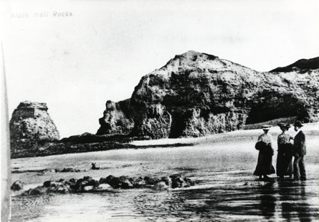 Postcard photograph entitled Black Hall Rocks, showing the cliffs with caves in them, the beach, and a man and a woman standing in the foreground
