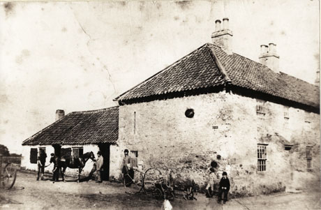 Photograph of the exterior of a cottage with a low one-storied building attached; outside the low building a horse is being shod with a man holding its head; another man is standing near the window of the cottage; two children are leaning on the wall of the cottage and a piece of agricultural machinery is lying on the ground in front of the children; the photograph is identified as Blacksmith's Shop, Castle Eden