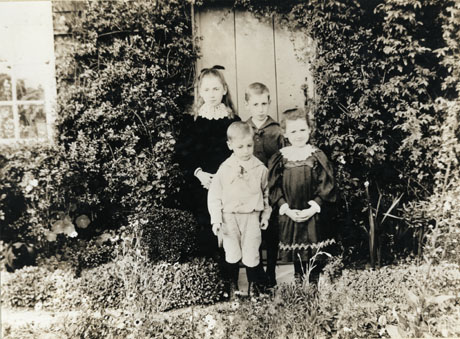Photograph of two small boys and two small girls, taken outside the door of a cottage the walls of which are covered with a creeper ; the children, who are approximately aged between four and ten years, are dressed in the clothes of the 1890s or 1900s, which can be seen clearly