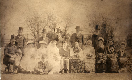 Photograph of a wedding group at Castle Eden showing the bride and groom and eight women and seven men in formal dress, photographed against a background of trees and fence