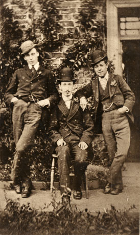 Photograph of three men dressed in smart suits outside a building in Castle Eden; one is sitting on a chair; all three are wearing button holes in their lapels