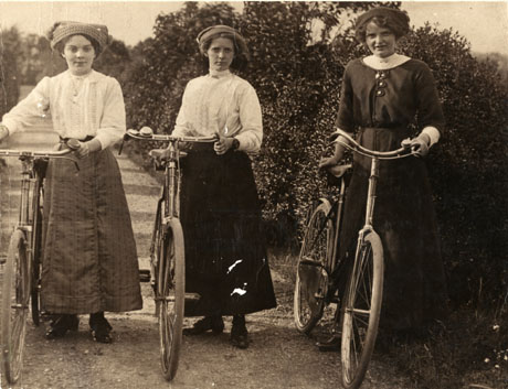 Photograph showing three young women facing the camera, dressed in ankle-length skirts, blouses and hats, standing next to, and holding by their handle bars, three bicycles; they are standing on a narrow road with a hedge down the right side; they have been identified as The Girls In Service, Castle Eden