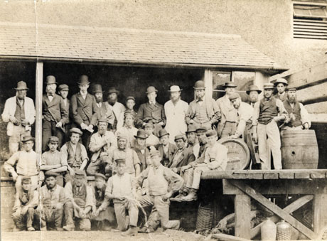 Photograph of forty five men and boys, posed in front of a platform with a roof, attached to a building; they are wearing suits, work clothes, caps, bowler, hats, waistcoats; they have two barrels, with the words Nimmo & Son, on them, near them on a platform on which some of the men are sitting; flasks in wickerwork can also be seen; they have been identified as employees of Nimmo's Brewery, Castle Eden