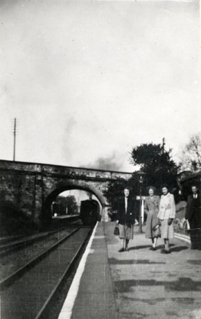 Photograph of three women and one man, identified as Fred Heads, Railways Area Manager, standing on the platform of a railway station, with, in the background, a railway bridge under which a train can be seen steaming away; the photograph is described as The Last Train To Leave Castle Eden
