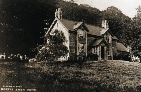 Postcard photograph entitled Garden of Eden Castle Eden Dene showing the exterior of the front of a substantial house backed by woodland; three indistinct figures can be seen at a table in the grounds of the house to the right of the picture