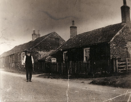 Photograph of the exterior of Ivy Cottage, back of the brewery, Castle Eden, with a number of similar low-roofed cottages in the distance; in front of the cottage is a man identified as a wheelwright and cartwright, whose blacksmith's shop was opposite the cottages shown and was on the site of the current golf club
