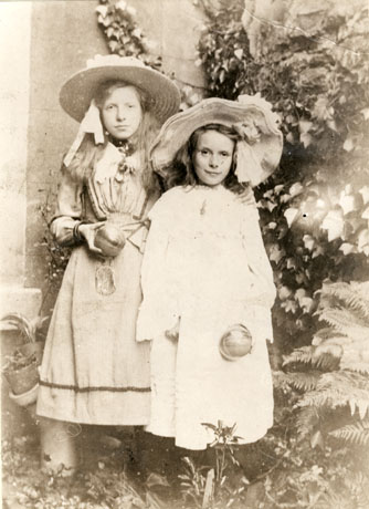 Photograph of two girls, of the ages of approximately ten and thirteen years, wearing elaborate dresses and hats and holding soft balls, described as coloured indoor balls, surrounded by what appear to be ferns, possibly in a photographer's studio
