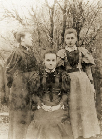 Photograph showing one young woman, dressed in an elaborate bodice and long skirt, sitting on a chair in the middle of the photograph looking at the camera; on the right, another young woman, similarly dressed, is standing looking at the camera; on the left, a third young woman, similarly dressed to the other two, is standing looking towards the young woman on the right; they have been identified as being in Castle Eden