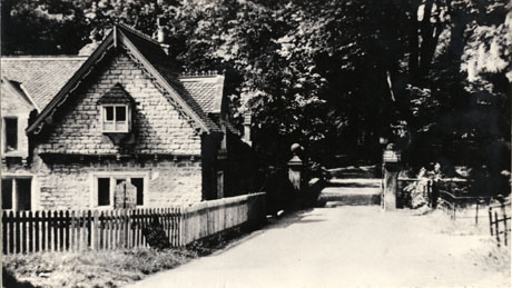 Photograph of the exterior of Dene Lodge with an extension at Castle Eden, showing a two-storied building surrounded by a low fence next to a road with pillars at the entrance to grounds filled with trees