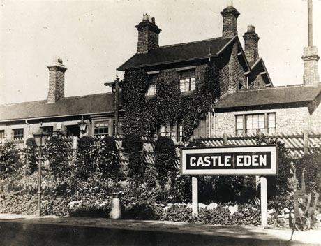 Photograph of the platform and buildings of Castle Eden Railway Station, showing the name of the station, flowers growing on the fence and, behind the fence, part of the facades of the buildings