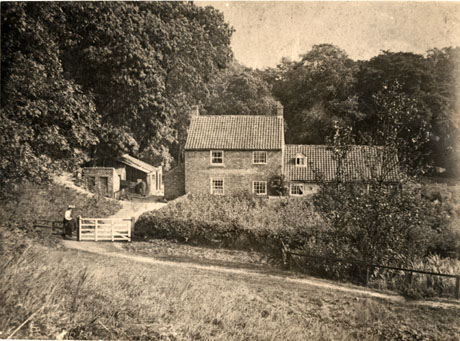 Photograph of the exterior of The Bleachery, Castle Eden, showing the front of a house of two stories, with a lower extension attached, surrounded by woods; in the foreground are a narrow track, a five-barred gate and an indistinct female figure sitting on a fence