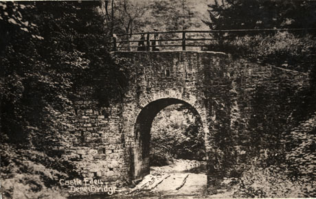 Postcard photograph entitled Castle Eden Dene Bridge, showing the bridge surrounded by woodland and with two indistinct female figures walking on the bridge, which crosses a road