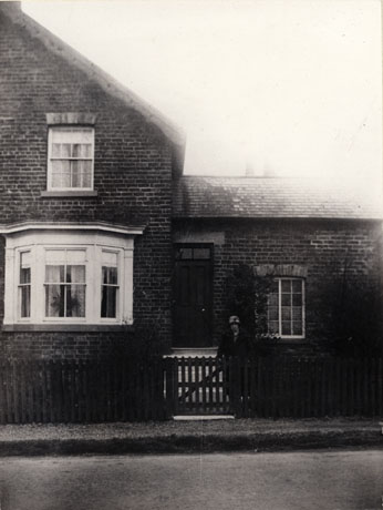 Photograph of the exterior of part of a house showing a bay window and a smaller window above it and part of a low building attached to the house; the building is described as The Ropery, Castle Eden; at the gate of the house is an indistinct female figure identified as Miss Elliott
