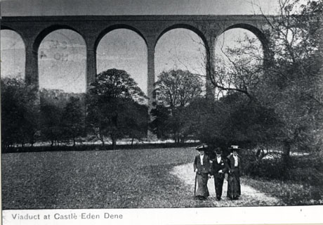 Postcard photograph entitled Viaduct at Castle Eden Dene showing, in the distance, six spans of the viaduct, with trees below it and, in the foreground, one man and two women walking in smart clothes