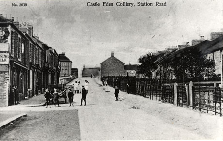 Postcard photograph entitled Castle Eden Colliery, Station Road, showing a road going uphill with terraced houses and shops on one side and houses set back from the road by their gardens on the other; a cart can be seen in the road near one of the shops; eight boys are standing in the foreground and three girls behind them in the middle distance; a few even more indistinct figures can be seen in the far distance; the photograph is also described as Station Road, Hesledon