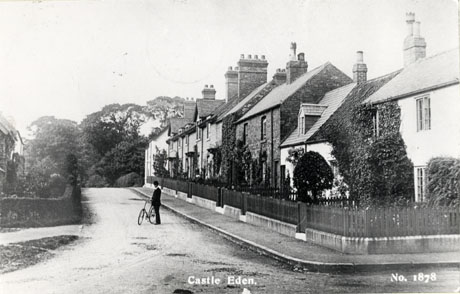 Postcard photograph entitled Castle Eden No 1878, showing one side of street of houses with small front gardens and creepers on the walls; only the gardens of the houses on the opposite side of the street can be seen; a man with a bicycle is standing in the middle of the road