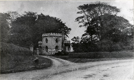 Photograph of the exterior of High Lodge, Castle Eden, with the lodge, a small castellated building near a gate, in the middle distance, and hedges and a road in the foreground