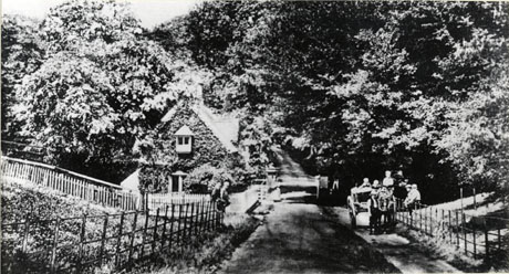 Photograph of The Lodge, Castle Eden taken from the same direction as cast0040, cast0046, cast0066, but from a greater distance; a cart may be seen on the right-hand side of the road with a number of indistinct figures near it; two indistinct figures are sitting on the fence on the opposite side of the road