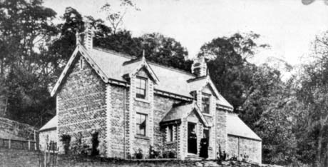 Photograph of the exterior of the side and front of a brick house with two dormer windows and a large porch with woodland behind and surrounding the house; the brickwork shows patterns at the corners of the house; two indistinct figures can be seen at the doorway in the porch; it has been described as Garden of Eden, Castle Eden