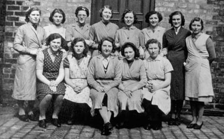 Photograph of fourteen women in overalls posed in front of a brick wall on a floor of brick; they have been identified as Nimmo's Brewery Staff, Castle Eden