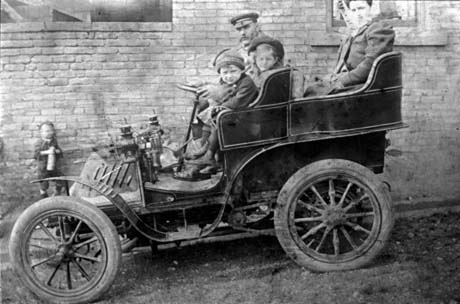 Photograph of the side view of an open car, with what appear to be solid wheels, on a slope in front of the wall of a house; a man is driving the car with two children on the front seat beside him; two women are sitting on the back seat; a blurred image of a small boy standing near the wall watching the car can be seen; the photograph has been described as An Outing, Castle Eden