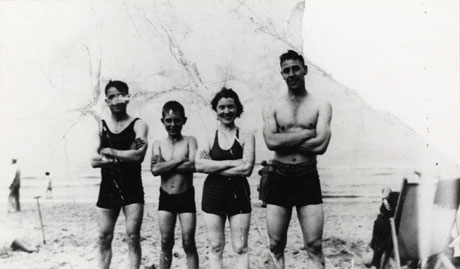 Photograph of two men, a woman, and a boy of about twelve years of age, in bathing costume, standing posed in a row, with the sea behind them; the top right-hand corner of the photograph is missing